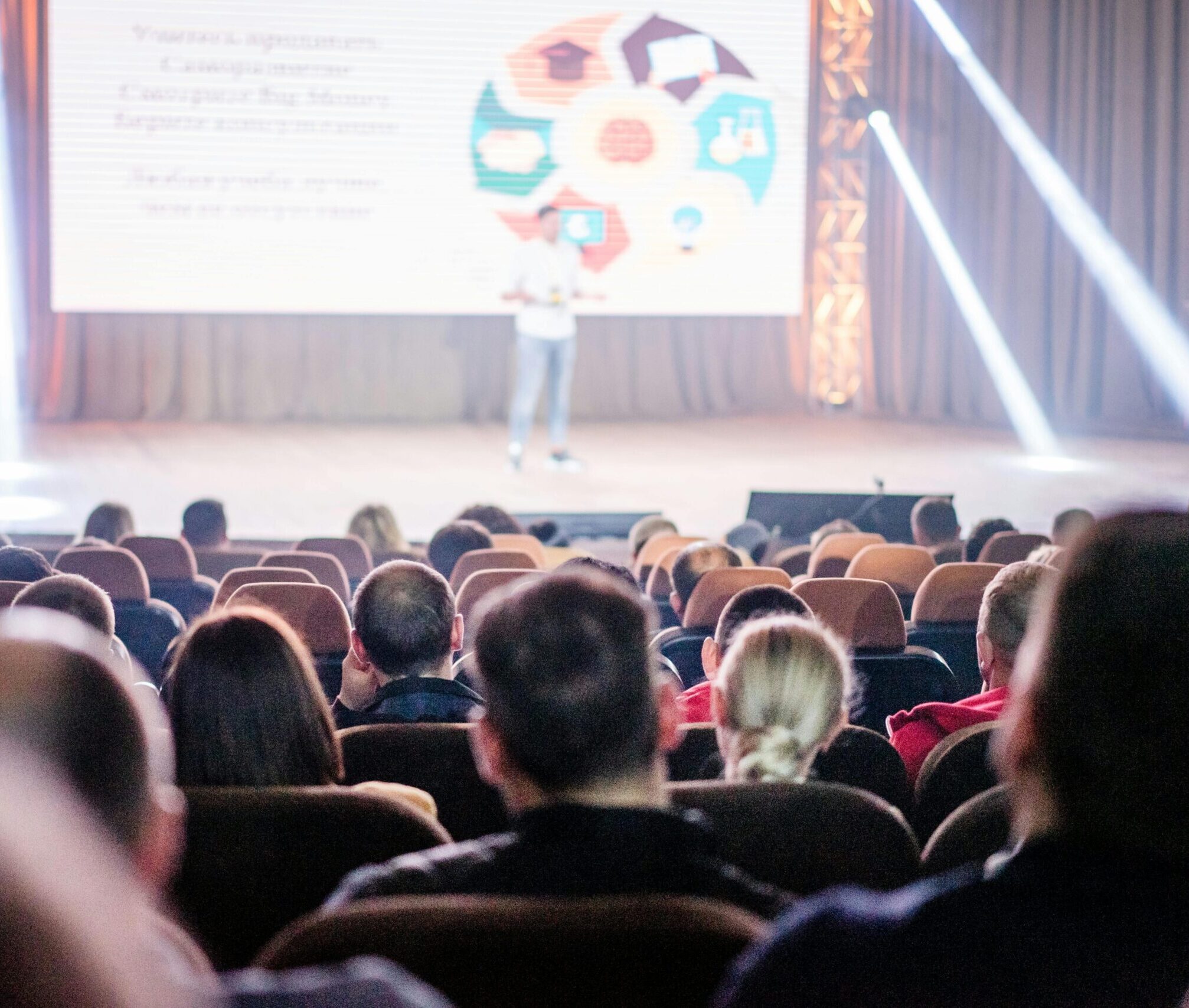 Audience attentively watching a presentation in a conference setting, with stage lights and speaker visible.
